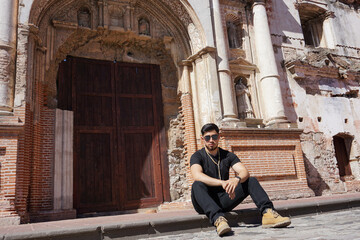 Fototapeta na wymiar Joven sentado afuera de una iglesia antigua pensando y reflexionando, serio y con mirada profunda