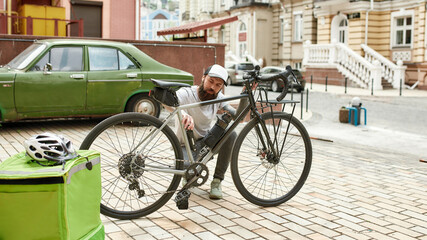 Naklejka na ściany i meble Be ready. Full length shot of brutal bearded delivery man in cap fixing, repairing his bicycle outdoors. Courier, delivery service concept