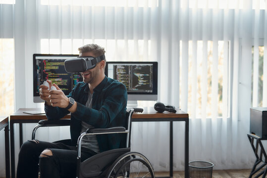 Having fun. Young male web developer in a wheelchair wearing virtual reality headset while working in the modern office. Multiple computer screens with program code. High Tech