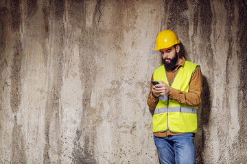 Young handsome bearded worker standing in front of concrete wall and using smart phone.