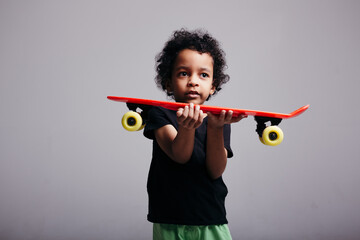A curly-skinned black boy holds out a red longboard and looks plaintively at the camera on a light background.