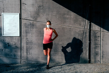 healthy caucasian woman in sportswear with protective mask stretching outdoor at sunset on a concrete wall background