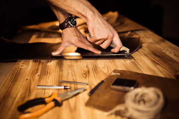 Working process of the leather belt in the leather workshop. Man holding crafting tool and working. Tanner in old tannery. Wooden table background. Close up man arm