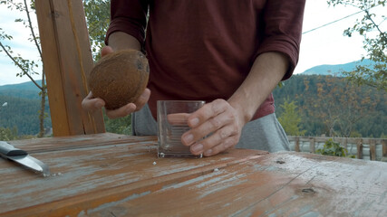 Man makes a hole in a coconut using knife, opens and pours coconut water into a transparent glass. Natural healthy organic tropical raw food. 