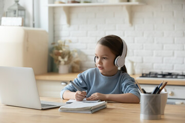 Concentrated schoolgirl sit at table in kitchen at home wear headphones learning using laptop app watching educational video makes notes. E-study, virtual easy convenient distant homeschooling concept