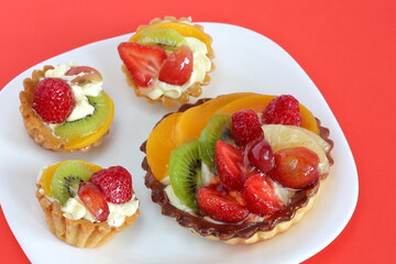Cupcake with strawberries, kiwi fruits, cherries, raspberries and peaches surrounded by strawberries, cut kiwi pieces on white plate against red background macro close up