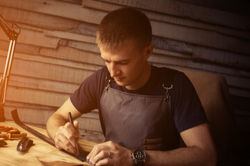 Fototapeta na wymiar Working process of the leather belt in the leather workshop. Man holding crafting tool and working. Tanner in old tannery. Wooden table background. Warm Light for text and design
