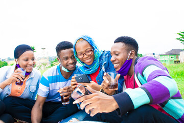 Obraz premium group of young black friends sitting outside together in a park, having drinks, watching content on a phone together, smiling and happy, wearing face masks