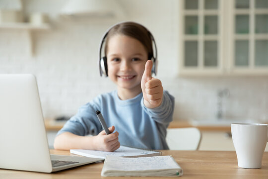 Close up focus on thumbs up of little girl learning at home wear headphones use pc smiles looks at camera. Easy foreign language e-learning, educational on-line app, self-education modern tech concept