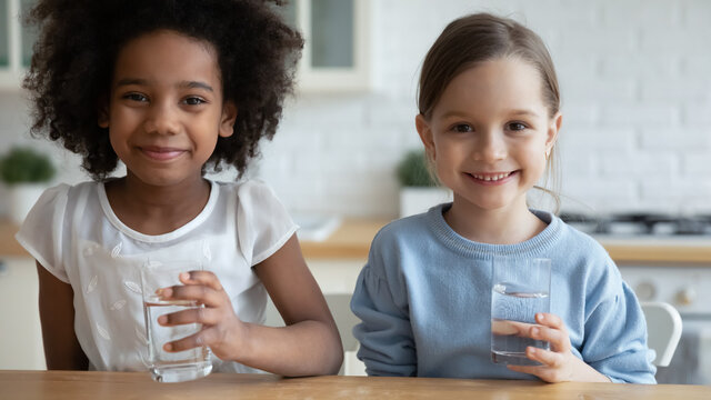 Close up horizontal image two school age girls african caucasian little adorable kids sit in kitchen holding glasses with still clear water look at camera. Healthy lifestyle, natural hydration concept