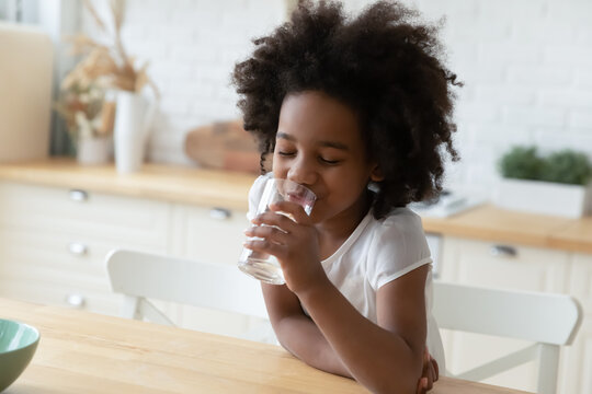 Close up image little mixed-race girl sitting at table in kitchen holding glass enjoy still or mineral water reduces thirst drinks clear aqua, healthy beverage, natural hydration body balance concept