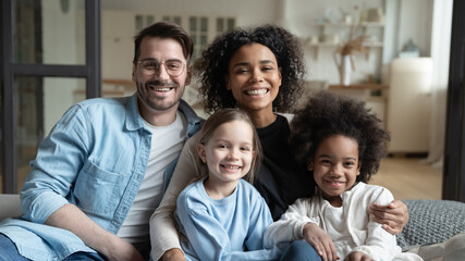 Multinational family married couple and daughters sit on couch looks at camera photo shooting feels...