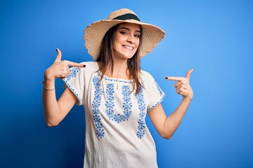 Young beautiful brunette woman wearing casual t-shirt and summer hat over blue background looking...