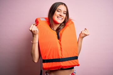 Young beautiful brunette woman wearing orange lifejacket over isolated pink background celebrating...