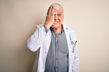 Senior handsome hoary doctor man wearing coat and stethoscope over white background covering one eye with hand, confident smile on face and surprise emotion.