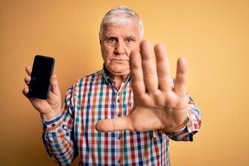 Senior handsome hoary man holding smartphone showing screen over yellow background with open hand doing stop sign with serious and confident expression, defense gesture