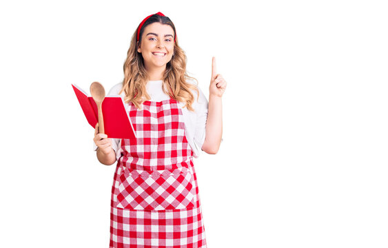 Young caucasian woman wearing apron holding recipe book and spoon surprised with an idea or question pointing finger with happy face, number one