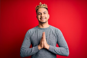 Young handsome man wearing golden crown of prince over isolated red background praying with hands...