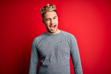 Young handsome man wearing golden crown of prince over isolated red background winking looking at...