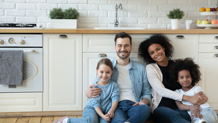 Horizontal photo multiracial excited couple with adorable daughters sit in modern renovated kitchen...