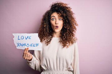 Young beautiful woman with curly hair and piercing holding paper with love yourself message scared in shock with a surprise face, afraid and excited with fear expression