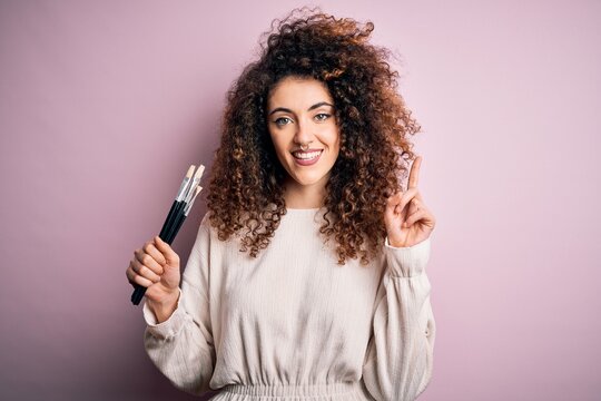 Young beautiful woman with curly hair and piercing applying cosmetic using paint brushes surprised with an idea or question pointing finger with happy face, number one