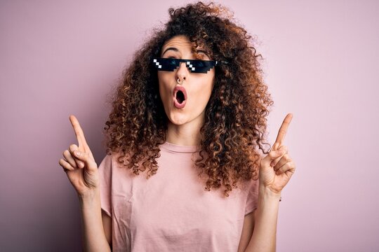 Young Beautiful Woman With Curly Hair And Piercing Wearing Funny Thug Life Sunglasses Amazed And Surprised Looking Up And Pointing With Fingers And Raised Arms.