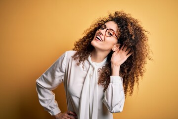 Young beautiful brunette woman with curly hair and piercing wearing shirt and glasses smiling with hand over ear listening an hearing to rumor or gossip. Deafness concept.