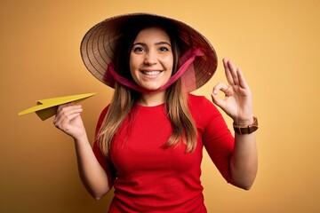 Tourist woman wearing traditional asian rice paddy straw hat holding paper plane for a trip doing ok sign with fingers, excellent symbol