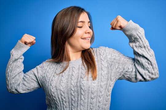 Beautiful young woman wearing casual wool sweater standing over blue isolated background showing arms muscles smiling proud. Fitness concept.