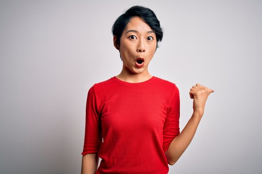 Young beautiful asian girl wearing casual red t-shirt standing over isolated white background Surprised pointing with hand finger to the side, open mouth amazed expression.