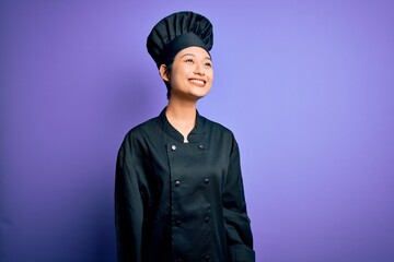 Young beautiful chinese chef woman wearing cooker uniform and hat over purple background looking away to side with smile on face, natural expression. Laughing confident.