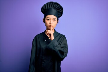 Young beautiful chinese chef woman wearing cooker uniform and hat over purple background asking to be quiet with finger on lips. Silence and secret concept.