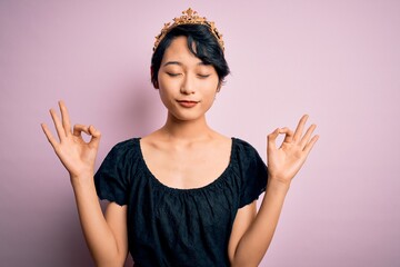 Young beautiful chinese woman wearing golden crown of king over isolated pink background relax and smiling with eyes closed doing meditation gesture with fingers. Yoga concept.