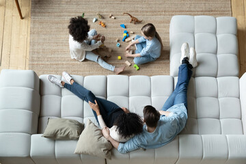 Above view parents sit on comfortable sofa while multi-ethnic daughters play on carpet using bricks...