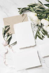 Wedding stationery, still life composition. Greeting cards mockup scene. Bouquet of white peony flowers, olive branches on marble, silk cloth. Vintage feminine photo, vertical flat lay, top view.
