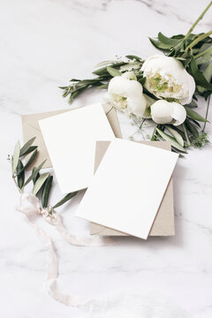 Wedding stationery, still life composition. Greeting cards mockup scene. Bouquet of white peony flowers, olive branches , cow parsley on marble, silk cloth. Vintage feminine photo, top view, defocused