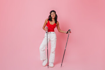 Glad young woman holding ski poles. Studio shot of trendy girl in sport pants.