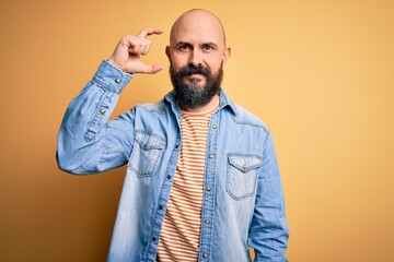Handsome bald man with beard wearing casual denim jacket and striped t-shirt smiling and confident gesturing with hand doing small size sign with fingers looking and the camera. Measure concept.