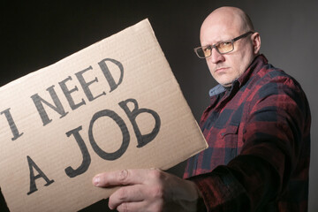 Man with a banner I need a job. Adult man in search of work. Concept - human lost his job. Concept - a man turned to the labor exchange. Bald human on a dark background. Protests of the unemployed.