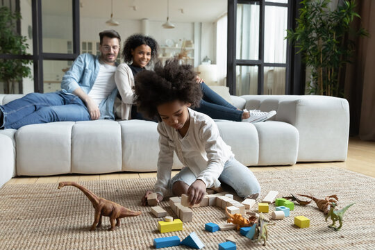 African Daughter Play On Floor With Wooden Blocks Set And Dinosaur Toys While Multi Ethnic Parents Sitting On Sofa In Living Room, Homeowners Spend Time At New Home. First House Family Weekend Concept