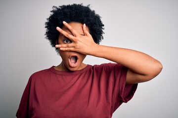 Fototapeta na wymiar Young beautiful African American afro woman with curly hair wearing casual t-shirt standing peeking in shock covering face and eyes with hand, looking through fingers with embarrassed expression.