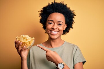 Young African American afro woman with curly hair holding bowl with chips potatoes very happy pointing with hand and finger