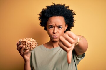 Young African American afro woman with curly hair holding bowl with salty peanuts with angry face,...