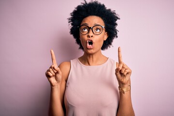 Young beautiful African American afro woman with curly hair wearing t-shirt and glasses amazed and surprised looking up and pointing with fingers and raised arms.