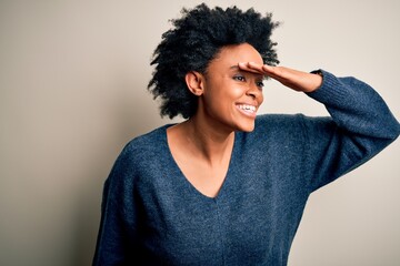 Fototapeta na wymiar Young beautiful African American afro woman with curly hair wearing casual sweater very happy and smiling looking far away with hand over head. Searching concept.