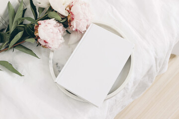 Wedding still life scene. White empty book cover mockup on marble tray. Pink peony flowers on white...