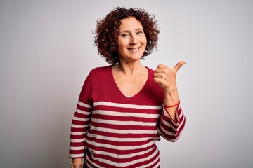 Middle age beautiful curly hair woman wearing casual striped sweater over white background smiling with happy face looking and pointing to the side with thumb up.