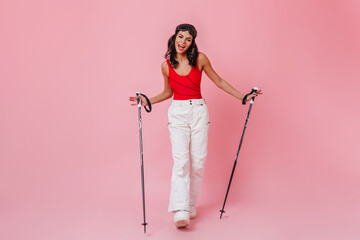 Charming tanned woman holding ski poles. Full length view of good-humoured girl in sport pants.