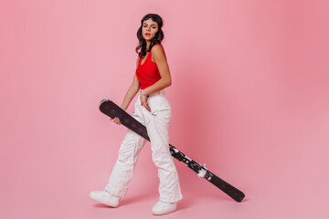 Cute young woman holding skis on pink background. Full length view of female model in sport goggles.